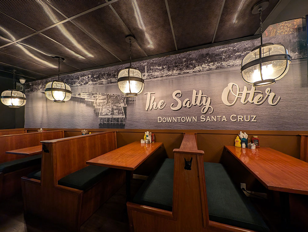 Wooden booths inside The Salty Otter with a large black-and-white Santa Cruz Wharf mural covering the wall.