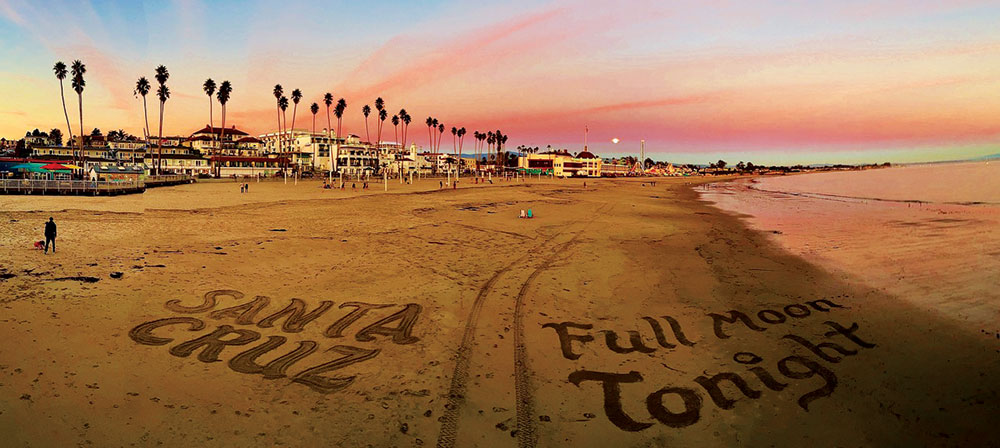 A panoramic sunset view of Capitola Beach with “Santa Cruz” and “Full Moon Tonight” written in large letters in the sand.