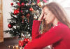 Holiday Hormones Woman in a red sweater looking stressed while sitting near a decorated Christmas tree.