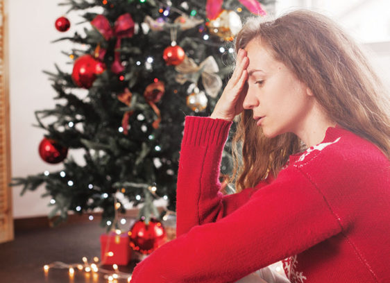 Holiday Hormones Woman in a red sweater looking stressed while sitting near a decorated Christmas tree.