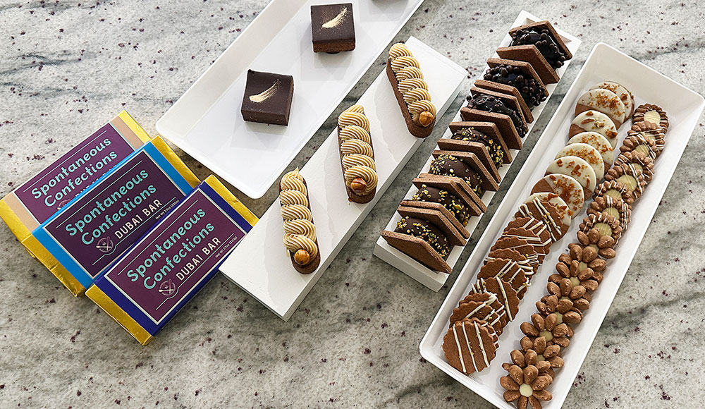 An assortment of Spontaneous Confections pastries and desserts arranged on white platters, including Dubai bars, brownies, tarts, and cookies.