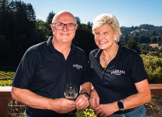 Chard Times Dave and Liz Ferrari of Ferrari Ranch Wines stand outdoors holding a glass of Chardonnay with vineyard hills in the background.