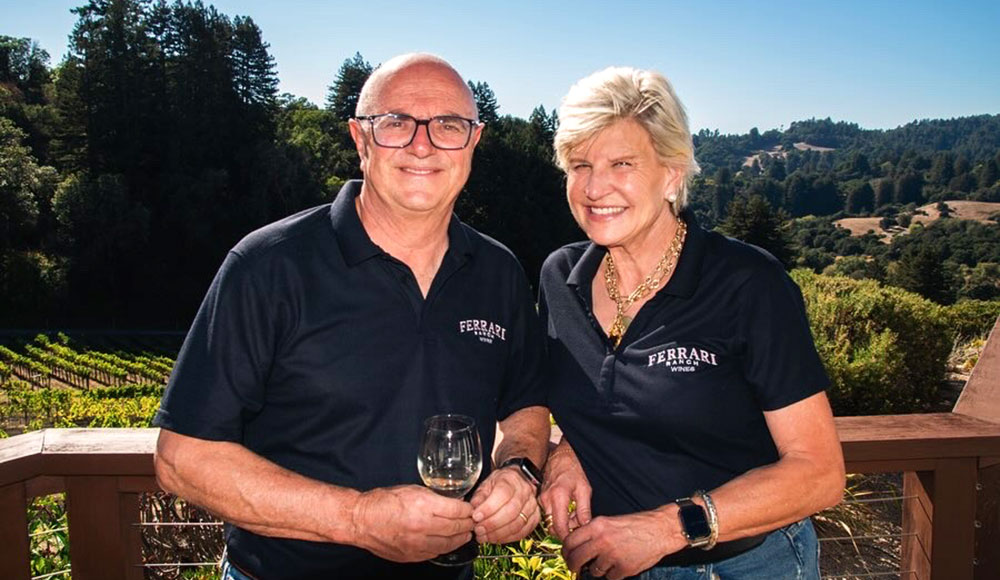 Dave and Liz Ferrari of Ferrari Ranch Wines stand outdoors holding a glass of Chardonnay with vineyard hills in the background.