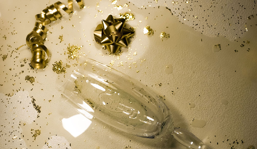 Broken champagne flute surrounded by gold ribbon and glitter on a white surface, symbolizing the aftermath of holiday overindulgence.
