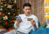 Scam Alert! Man sitting on a couch near a Christmas tree looks concerned while checking a credit card and smartphone.