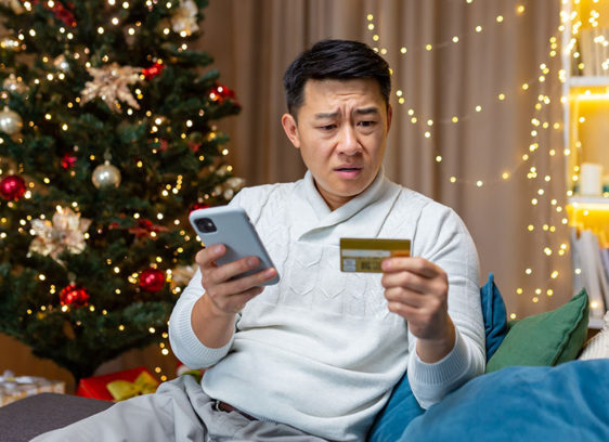 Scam Alert! Man sitting on a couch near a Christmas tree looks concerned while checking a credit card and smartphone.