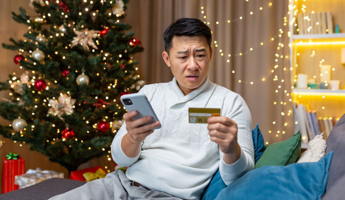 Scam Alert! Man sitting on a couch near a Christmas tree looks concerned while checking a credit card and smartphone.