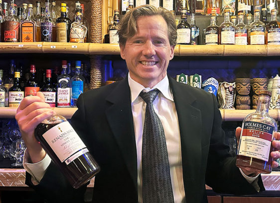 Santa Cruz’s Rum Baron Man holding two bottles behind a bar in front of a shelf full of liquor