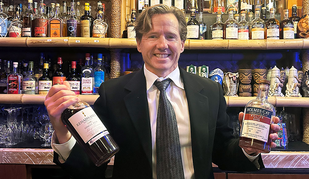 Man holding two bottles behind a bar in front of a shelf full of liquor