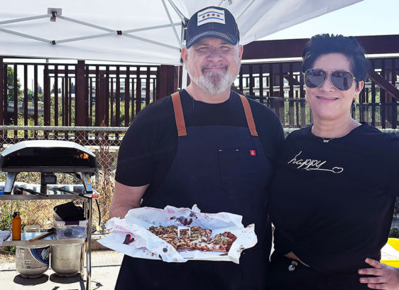 Sunflower Soul J.R. Chapman and Viviana of Girasol Pizza hold a Chicago-style thin-crust pizza at a pop-up food setup.