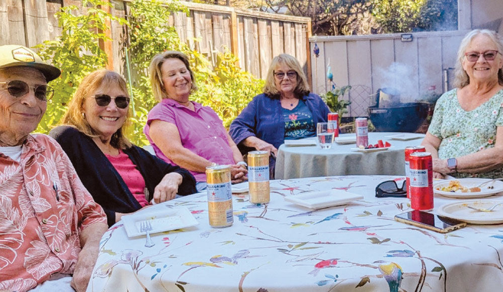 Members of Village Santa Cruz County gather around a table outdoors, enjoying conversation and refreshments together.