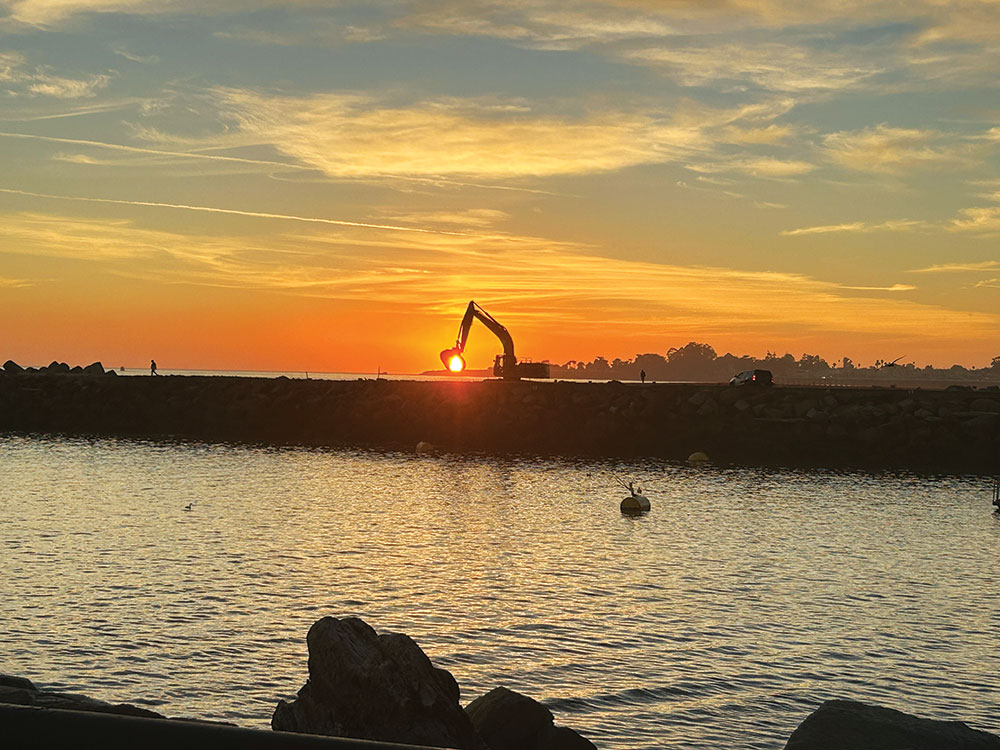 Sunset over Santa Cruz Harbor with a silhouetted excavator on the breakwater and calm water in the foreground.