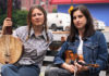 Strings Attached Musicians Nora Brown and Stephanie Coleman sit on a bench holding a banjo and violin in an outdoor setting.
