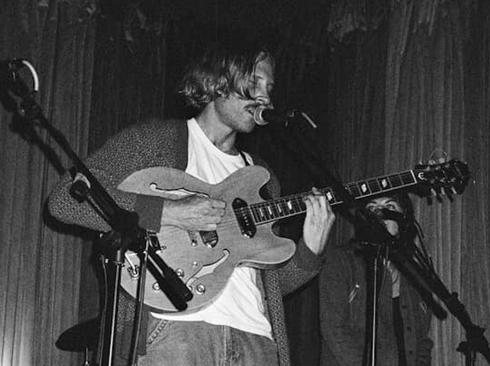 Musician Richard Tripps sings into a microphone while playing an electric guitar onstage in a black-and-white photo.