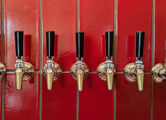 Level-Up Liquids A row of polished beer taps mounted against a glossy red tile wall inside Other Brother Beer Co.