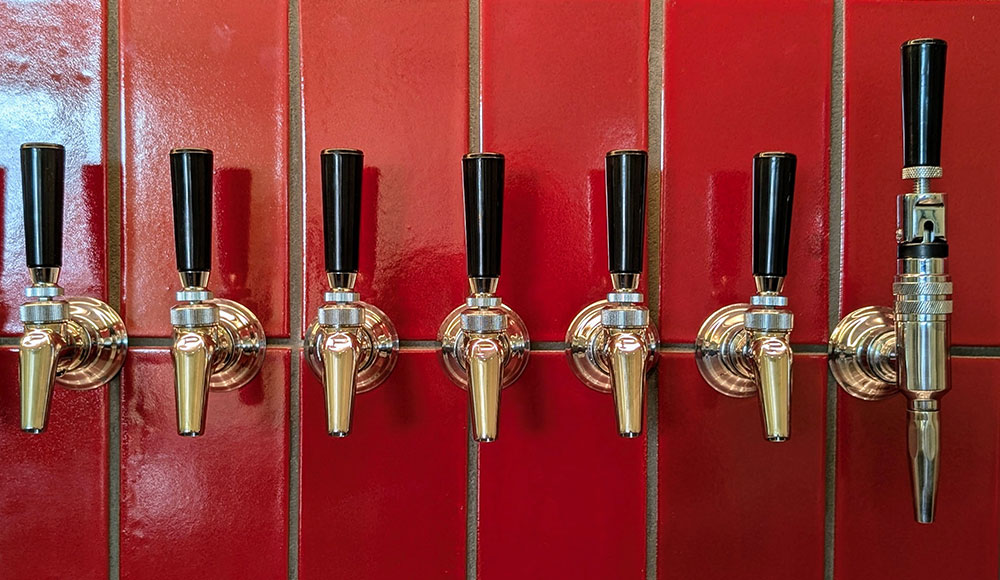 A row of polished beer taps mounted against a glossy red tile wall inside Other Brother Beer Co.