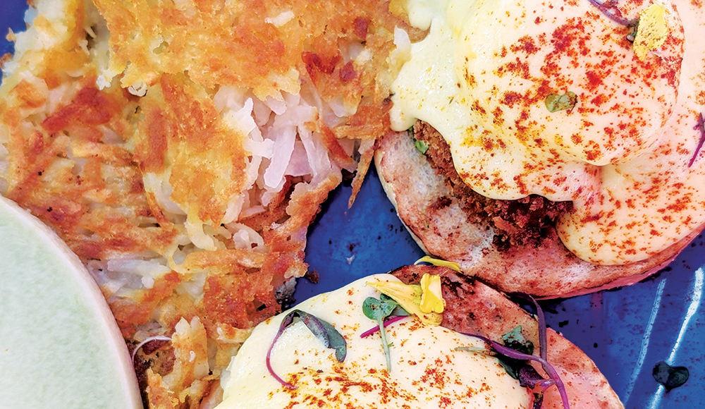 Crab cake Benedict served with poached eggs, hollandaise sauce, and crispy hash browns on a blue plate.