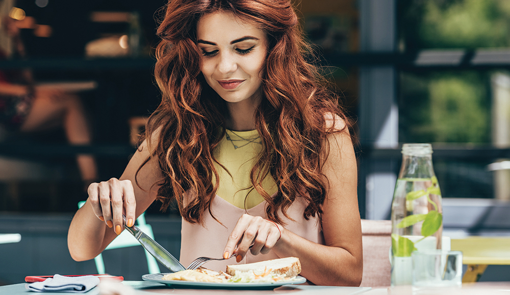 A woman sits at a table cutting food on a plate during a solo meal.