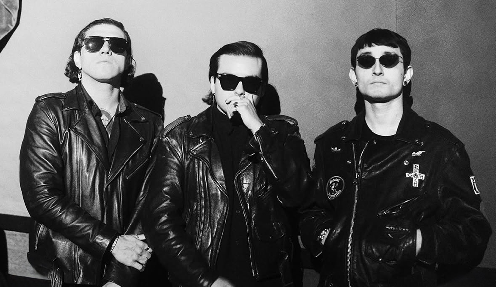 Three musicians wearing black leather jackets and sunglasses pose for a black-and-white portrait.