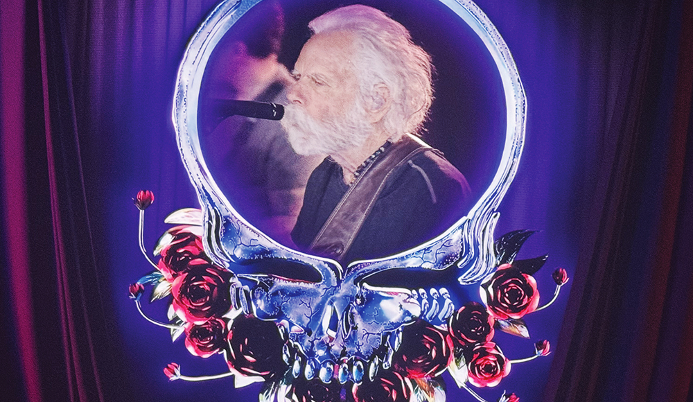 An older Bob Weir sings into a microphone, framed by a glowing skull-and-roses stage graphic.