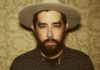 Welcome to the Family Musician Jackie Greene poses for a portrait wearing a hat against a patterned background.