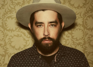 Welcome to the Family Musician Jackie Greene poses for a portrait wearing a hat against a patterned background.