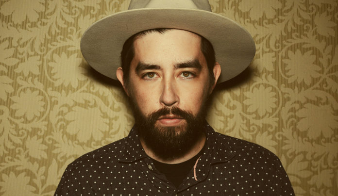 Welcome to the Family Musician Jackie Greene poses for a portrait wearing a hat against a patterned background.