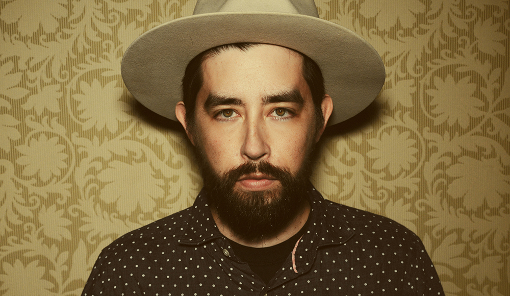 Musician Jackie Greene poses for a portrait wearing a hat against a patterned background.