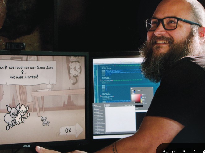 Edmund McMillen sits at a desk smiling beside a computer screen displaying artwork from a video game in development.