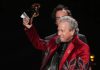 The Editor’s Desk John Leopold in a red suit holds up a Grammy Award while speaking at a podium onstage