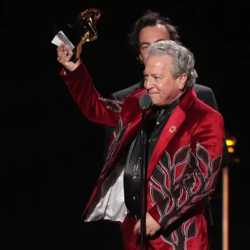 John Leopold in a red suit holds up a Grammy Award while speaking at a podium onstage