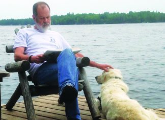 Books of the Dead A man sits on a wooden dock reading a book while petting a light-colored dog beside a lake.