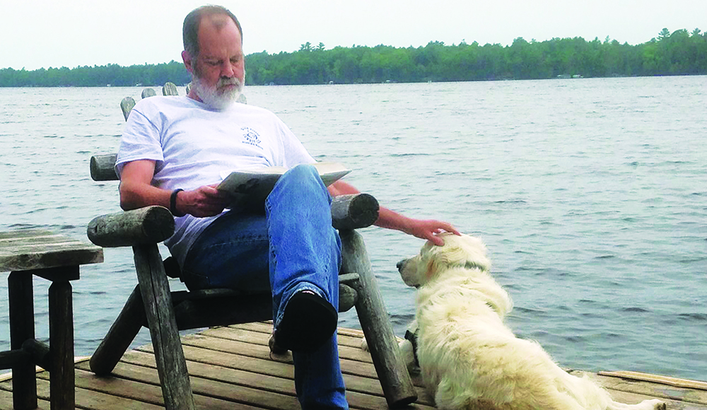 A man sits on a wooden dock reading a book while petting a light-colored dog beside a lake.
