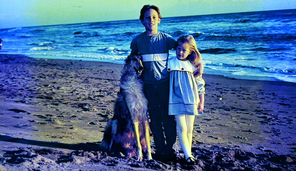 Two children stand on a beach with a dog near the shoreline