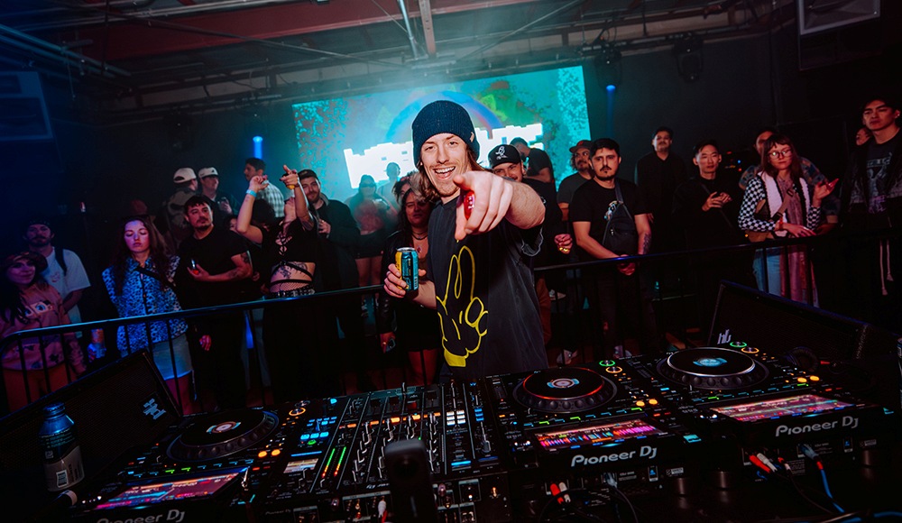 DJ performing live at a crowded Santa Cruz nightclub, pointing toward the camera behind a Pioneer DJ mixing console