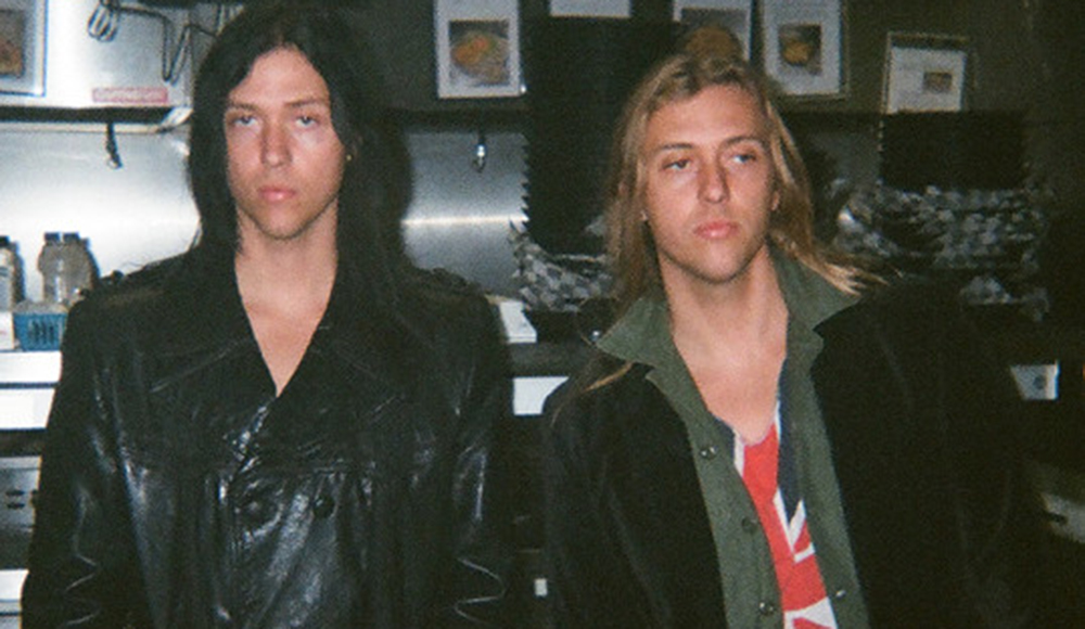Two musicians standing indoors with long hair and leather jackets