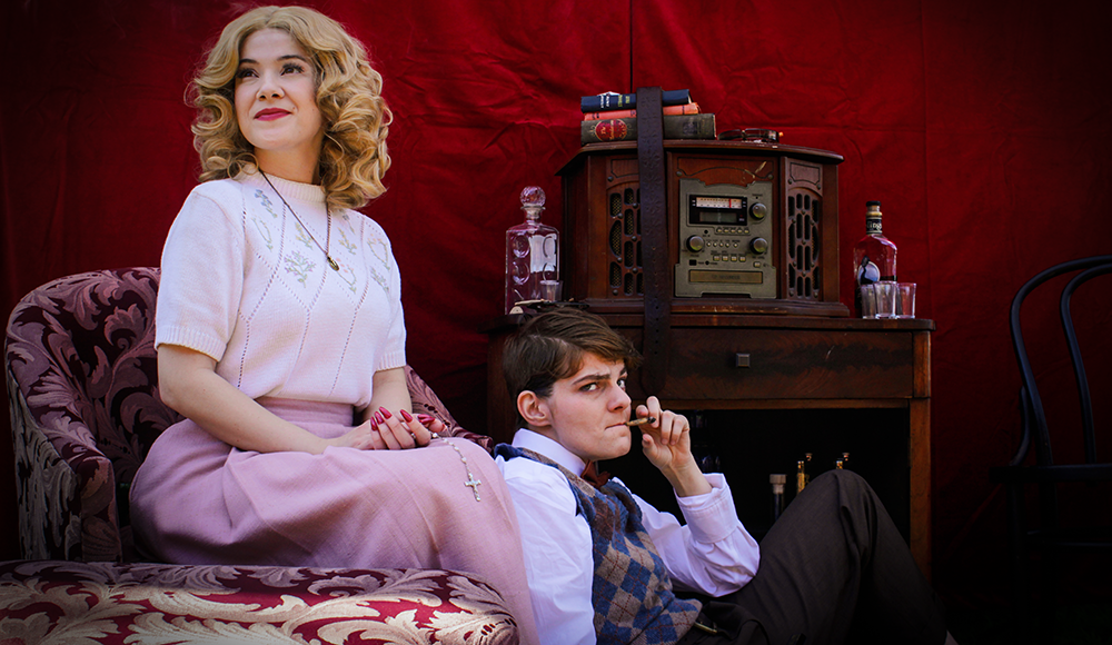 Two actors in vintage clothing seated near old record player against red background
