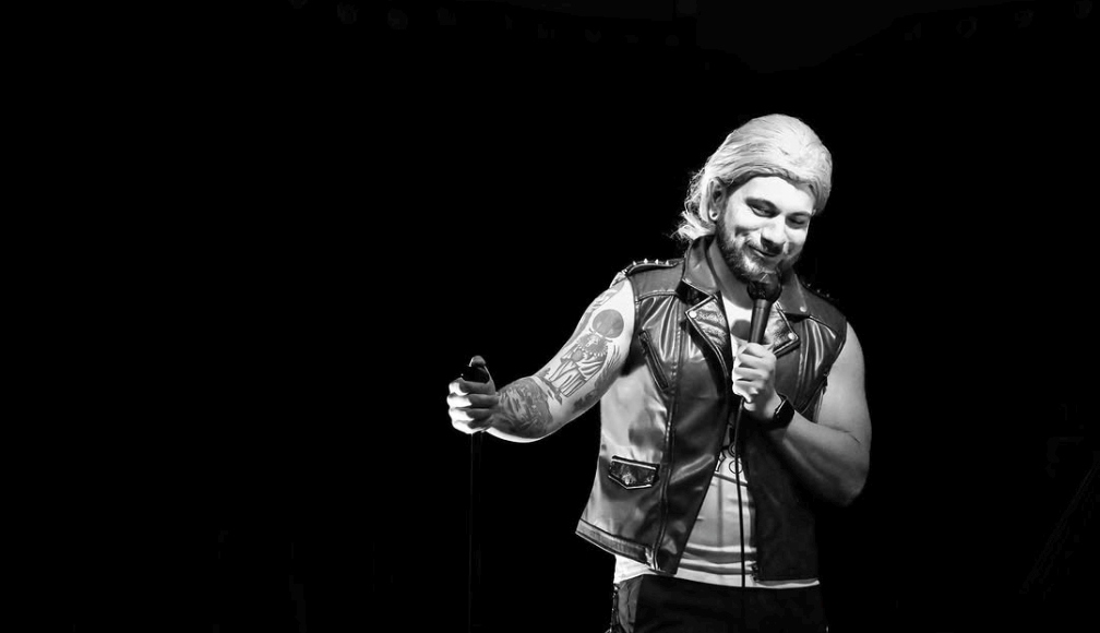 Black-and-white photo of a stand-up comedian in a leather vest holding a microphone on stage at Blue Lagoon in Santa Cruz