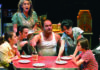 He Escaped Queens Cast members gather around a red kitchen table in a scene from Joe Ortiz’s musical Escaping Queens.