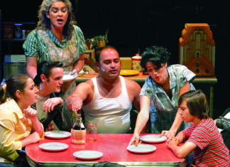 He Escaped Queens Cast members gather around a red kitchen table in a scene from Joe Ortiz’s musical Escaping Queens.