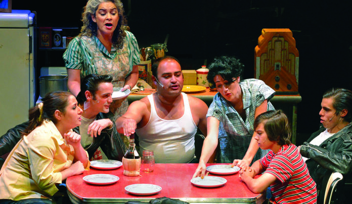 He Escaped Queens Cast members gather around a red kitchen table in a scene from Joe Ortizโs musical Escaping Queens.