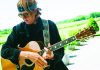 Music and Art Brett Dennen holds an acoustic guitar outdoors in a sunny vineyard setting.