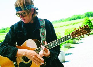 Music and Art Brett Dennen holds an acoustic guitar outdoors in a sunny vineyard setting.
