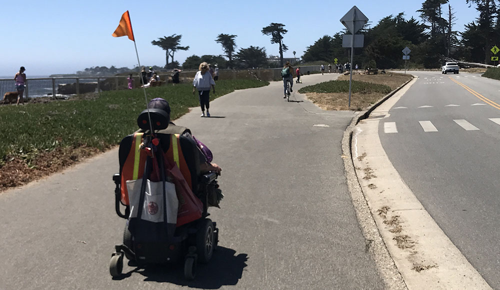 A person using a mobility scooter travels along the West Cliff Drive path in Santa Cruz, separated from car traffic.