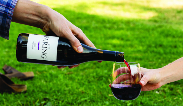 Crimson & Clover A bottle of The Paring red wine being poured into a glass outdoors on a green lawn.