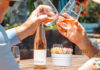 Summer Swirls Two people toast glasses of Bouchaine Vin Gris of Pinot Noir beside a bottle on an outdoor table.