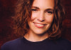No Glass Ceiling Comedian Beth Stelling smiles in a close-up portrait against a dark red background.