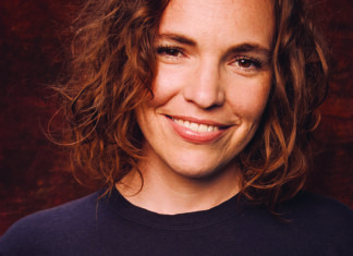 No Glass Ceiling Comedian Beth Stelling smiles in a close-up portrait against a dark red background.