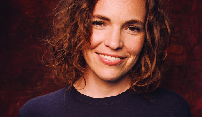 No Glass Ceiling Comedian Beth Stelling smiles in a close-up portrait against a dark red background.