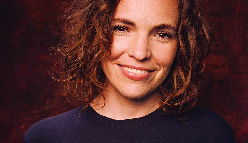 Comedian Beth Stelling smiles in a close-up portrait against a dark red background.